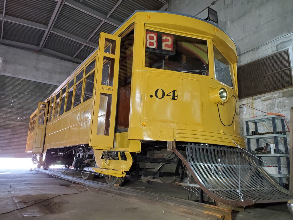 Mostly restored .04 trolley; the yellow paint is bright, the body repaired, door replaced, and a cow-catcher guard affixed to the front.